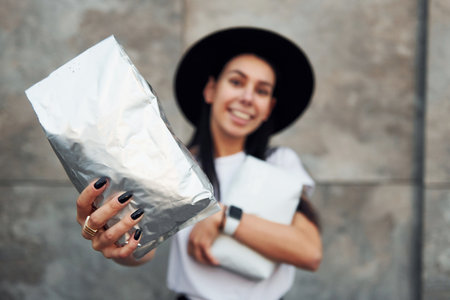 Brunette in black hat and white shirt standing near the grey wall and holds package with coffeeの写真素材