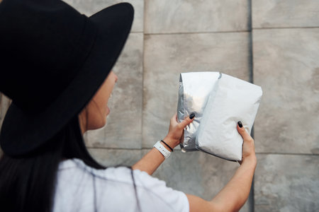 Brunette in black hat and white shirt standing near the grey wall and holds package with coffeeの写真素材