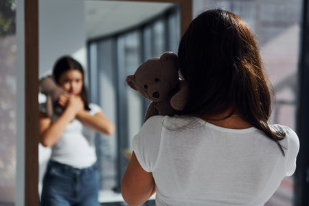 Young woman holds teddy bear and looks at herself into the mirrorの写真素材