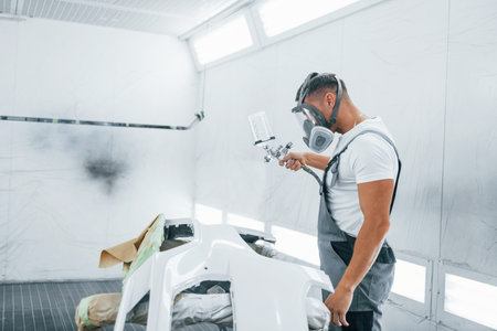 Uses painting gun. Caucasian automobile repairman in uniform works in garageの写真素材