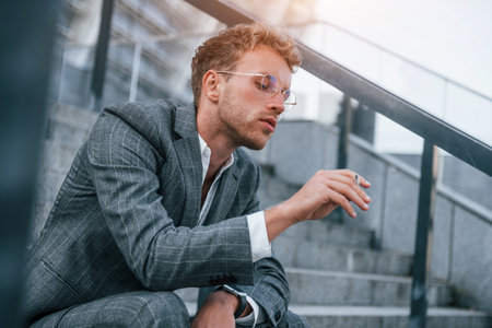 Smoking cigarette. Young businessman in grey formal wear is outdoors in the cityの写真素材