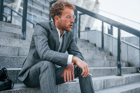 Smoking cigarette. Young businessman in grey formal wear is outdoors in the cityの写真素材