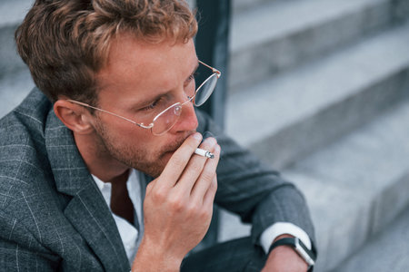 Takes a break and smokes a cigarette. Young businessman in grey formal wear is outdoors in the cityの写真素材