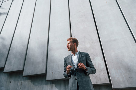 Standing against building wall. Young businessman in grey formal wear is outdoors in the cityの写真素材