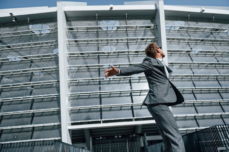 Dancing, celebrating success. Young businessman in grey formal wear is outdoors in the cityの写真素材