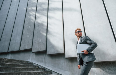 Runs and lating to the job. Young businessman in grey formal wear is outdoors in the cityの写真素材