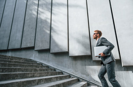 Runs and lating to the job. Young businessman in grey formal wear is outdoors in the cityの写真素材