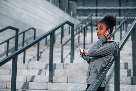 Listens to music in headphones. Young african american woman in sportive clothes have workout outdoors at daytimeの写真素材