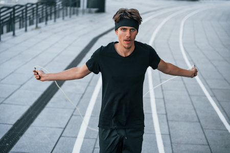 Uses jumping rope. Young man in sportive clothes have workout outdoors at daytimeの写真素材