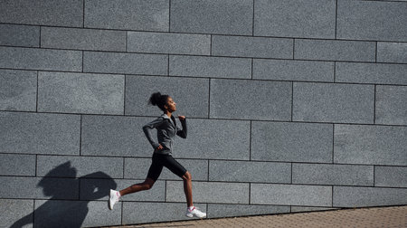 Side view of young african american woman in sportive clothes that runs near wallの写真素材