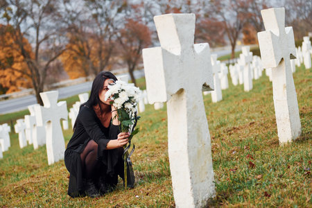 With flowers in hands. Young woman in black clothes visiting cemetery with many white crosses. Conception of funeral and deathの写真素材