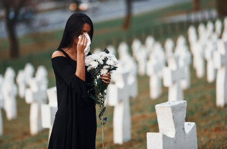 Young woman in black clothes visiting cemetery with many white crosses. Conception of funeral and deathの写真素材