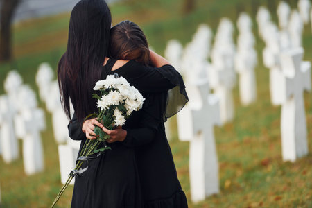 Embracing each other and crying. Two young women in black clothes visiting cemetery with many white crosses. Conception of funeral and deathの写真素材
