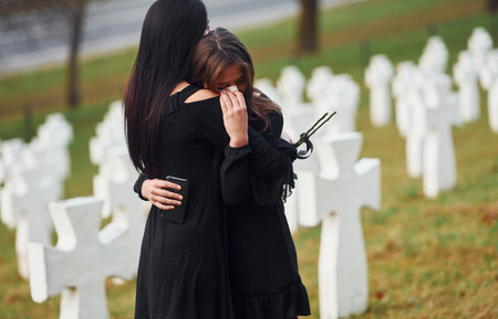 Embracing each other and crying. Two young women in black clothes visiting cemetery with many white crosses. Conception of funeral and deathの写真素材