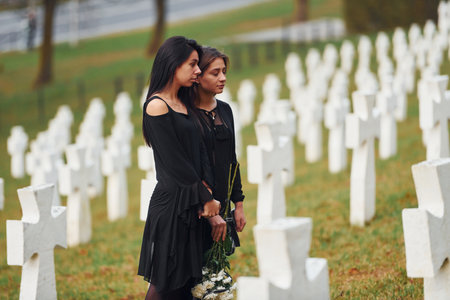 Holds flowers. Two young women in black clothes visiting cemetery with many white crosses. Conception of funeral and deathの写真素材