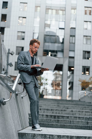 Laptop in hands. Young elegant man in good clothes is outdoors in the city at daytimeの写真素材