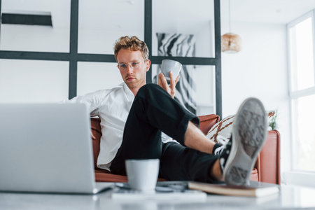 With laptop. Caucasian young guy in elegant white shirt indoors at home