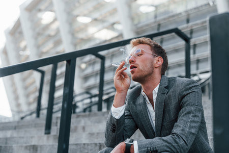Sits and smokes cigarette. Young successful businessman in grey formal wear is outdoors in the cityの写真素材