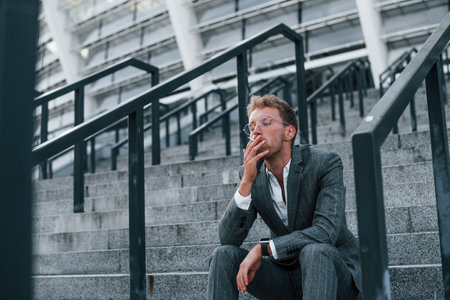 Sits and smokes cigarette. Young successful businessman in grey formal wear is outdoors in the cityの写真素材