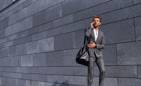 Talks by phone. Young successful businessman in grey formal wear is outdoors in the cityの写真素材