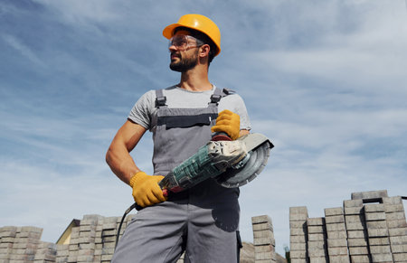 Posing for a camera with circular saw. Male worker in yellow colored uniform have job with pavementの写真素材