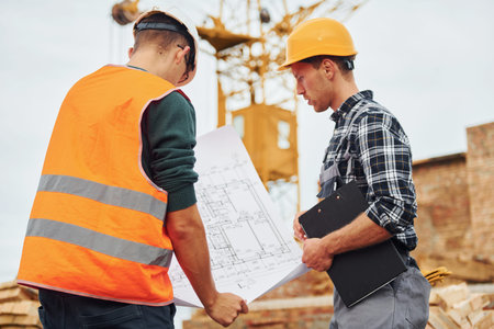 Using paper document with plan on it. Two construction workers in uniform and safety equipment have...