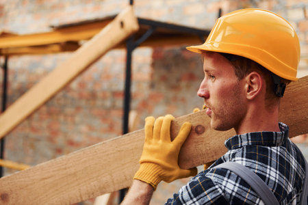 Side view of man that holds wooden boards. Construction worker in uniform and safety equipment have job on buildingの写真素材