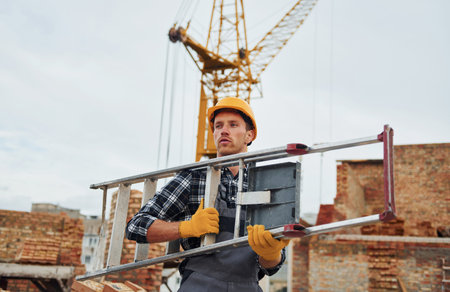Ladder in hands. Construction worker in uniform and safety equipment have job on buildingの写真素材