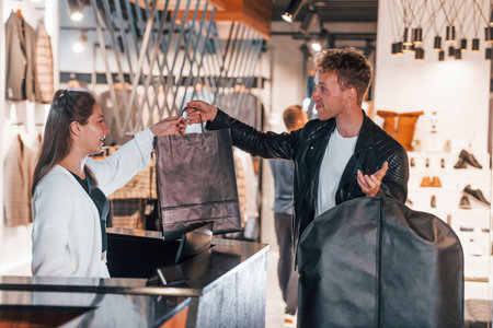 Man makes purchase in modern store with elegant clothes. Female worker serves the guy at the checkoutの写真素材