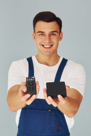 TV remote controller and smartphone. Male worker in blue uniform standing inside of studio against...