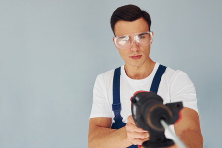 In protective eyewear and with drill in hands. Male worker in blue uniform standing inside of studio against white backgroundの写真素材