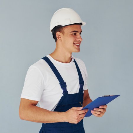 With notepad and hard hat. Male worker in blue uniform standing inside of studio against white backgroundの写真素材