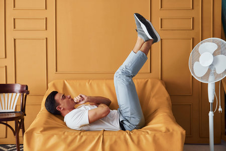Young guy with stylish haircut and in casual clothes sitting indoors in room with orange colored...
