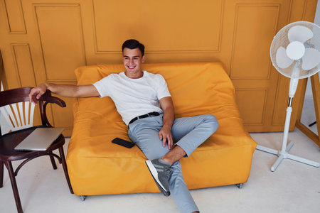 Young guy with stylish haircut and in casual clothes sitting indoors in room with orange colored interiorの写真素材