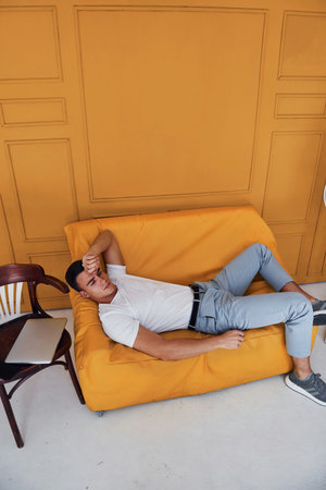 Young guy with stylish haircut and in casual clothes sitting indoors in room with orange colored interiorの写真素材