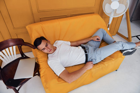 Young guy with stylish haircut and in casual clothes sitting indoors in room with orange colored...