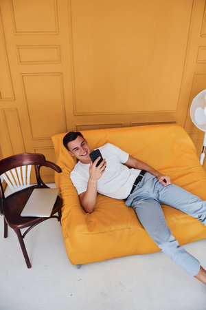 Young guy with stylish haircut and in casual clothes sitting indoors in room with orange colored interiorの写真素材