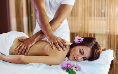 Purple flower lying down. Man does massage to the young woman in white towel indoorsの写真素材