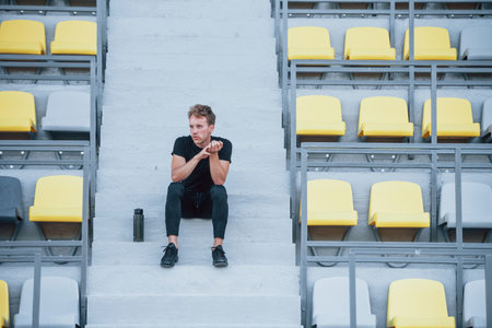 Sits with bottle of water. Sportive young guy in black shirt and pants outdoors at daytimeの写真素材