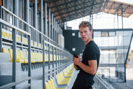 Standing and holding bottle of water. Sportive young guy in black shirt and pants outdoors at daytimeの写真素材