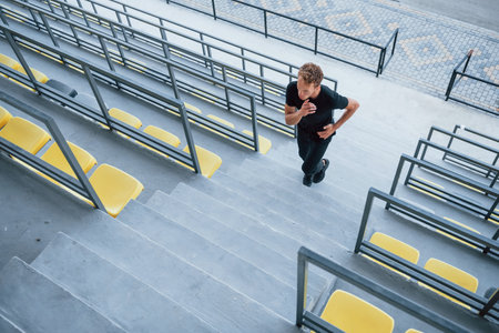 Running on the bleachers. Sportive young guy in black shirt and pants outdoors at daytimeの写真素材