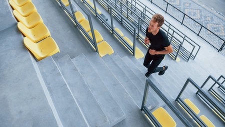 Running on the bleachers. Sportive young guy in black shirt and pants outdoors at daytime