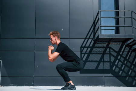 Exercising near building with black wall. Sportive young guy in black shirt and pants outdoors at daytimeの写真素材