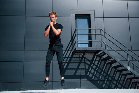 Exercising near building with black wall. Sportive young guy in black shirt and pants outdoors at daytimeの写真素材