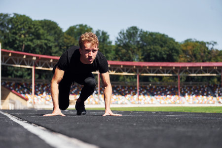 Runner is on starting position on track. Sportive young guy in black shirt and pants outdoors at daytimeの写真素材