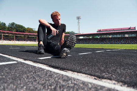 Tired runner sits on track and taking a break. Sportive young guy in black shirt and pants outdoors at daytimeの写真素材
