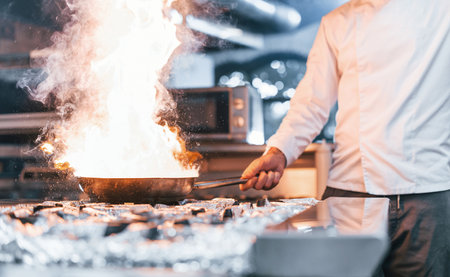 Frying pan is on fire. Chef in white uniform cooking food at kitchen. Busy day at workの写真素材