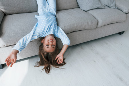 Lying upside down on the sofa. Cute little girl indoors at home alone. Child enjoying weekendの写真素材