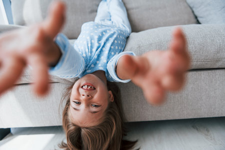 Lying upside down on the sofa. Cute little girl indoors at home alone. Child enjoying weekendの写真素材