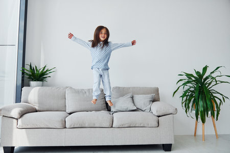 Jumping on the sofa. Cute little girl indoors at home alone. Child enjoying weekendの写真素材